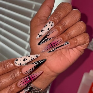 Puede incluir: Uñas acrílicas largas y puntiagudas con varios diseños. Algunas uñas son blancas con lunares negros, otras tienen puntas negras con puntos blancos y algunas presentan patrones de puntos rosas y negros. Se ve una pulsera plateada.