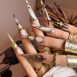 May include: Long, pointed acrylic nails with gold accents and religious-themed charms. The nails feature a variety of designs, including gold crosses, gold beads, and marble-like patterns in white and brown. Gold rings adorn the fingers.