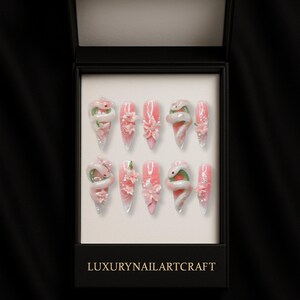 May include: A set of ten artificial nails in a black box. The nails are pink with white and green snake and flower designs. The box has the text "LUXURYNAILARTCRAFT" in gold lettering.