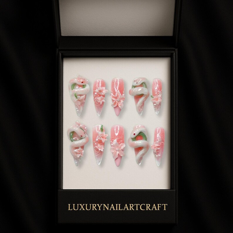 May include: A set of ten artificial nails in a black box. The nails are pink with white and green snake and flower designs. The box has the text "LUXURYNAILARTCRAFT" in gold lettering.