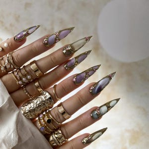 May include: A hand displaying long, ornate, stiletto-shaped nails. Each nail features a gold-toned metal design with lavender, pearl, and pink accents. The fingers are adorned with multiple gold rings of various styles.