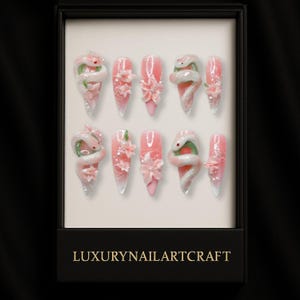 May include: A set of ten artificial nails in a black box. The nails are pink with white and green snake and flower designs. The box has the text "LUXURYNAILARTCRAFT" in gold lettering.