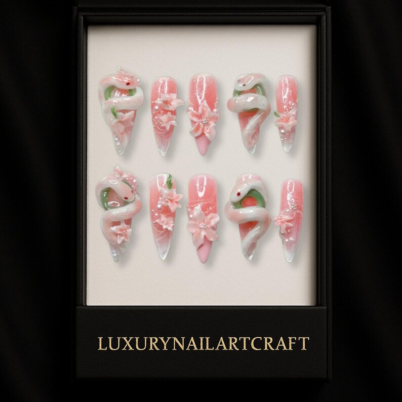 May include: A set of ten artificial nails in a black box. The nails are pink with white and green snake and flower designs. The box has the text "LUXURYNAILARTCRAFT" in gold lettering.