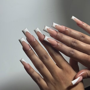 May include: A close-up of a hand with long, square-shaped acrylic nails. The nails feature a classic French manicure design with a nude base and a thick white tip. The nails are well-manicured and glossy.