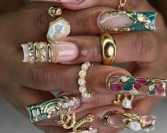 May include: Long square-shaped artificial nails featuring intricate gold metallic swirls, embedded white pearls, and multi-colored translucent panels. The nails are adorned with various gold rings, including a snake design, a smooth band, and a pearl-encrusted ring.