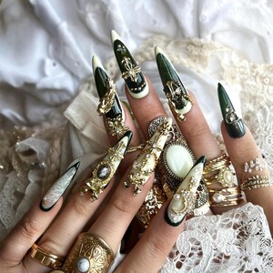 Puede incluir: Primer plano de una mano con u&ntilde;as largas y puntiagudas, elaboradamente decoradas. Las u&ntilde;as presentan dise&ntilde;os en verde oscuro, crema y blanco, adornadas con adornos dorados, joyas y perlas. Varios anillos dorados adornan los dedos.