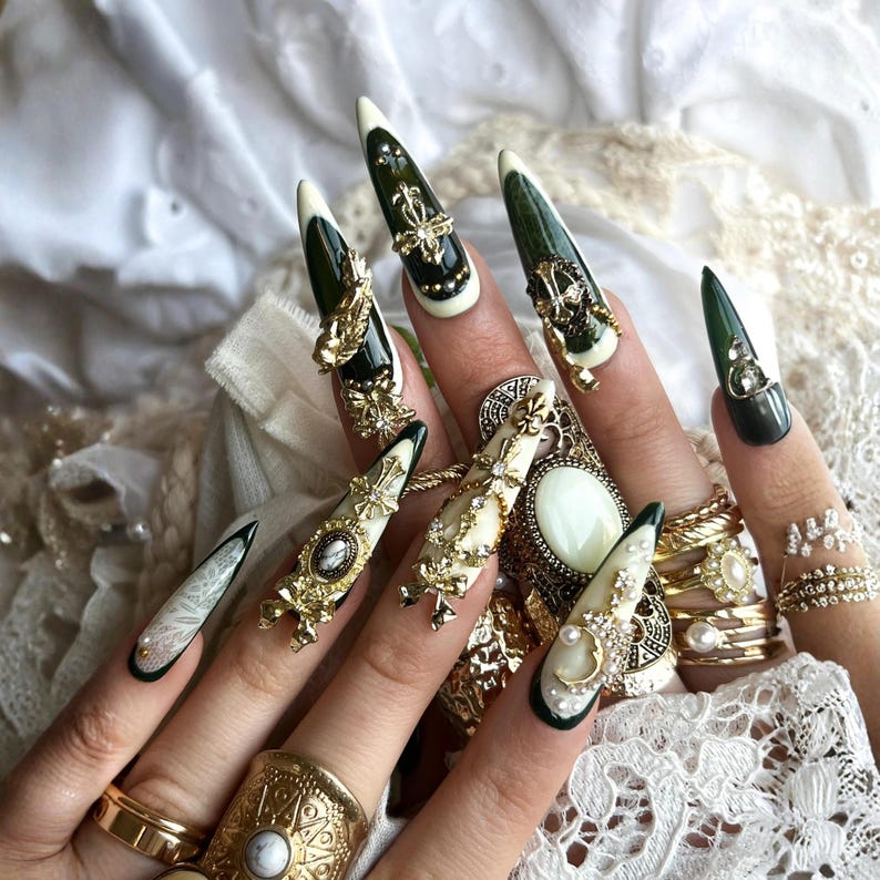 Puede incluir: Primer plano de una mano con u&ntilde;as largas y puntiagudas, elaboradamente decoradas. Las u&ntilde;as presentan dise&ntilde;os en verde oscuro, crema y blanco, adornadas con adornos dorados, joyas y perlas. Varios anillos dorados adornan los dedos.