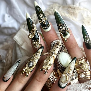 Puede incluir: Una mano que muestra u&ntilde;as artificiales largas, puntiagudas y elaboradamente decoradas. Las u&ntilde;as presentan una base verde oscuro con puntas blancas, adornadas con adornos dorados, joyas y dise&ntilde;os ornamentados. Tambi&eacute;n se ven varios anillos dorados.