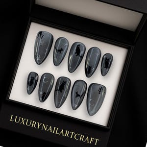 May include: A set of glossy, dark gray press-on nails with a black cat and celestial design. The nails are almond-shaped and arranged in a black box with the text "LUXURY NAILARTCRAFT". The nail art features moons and stars.