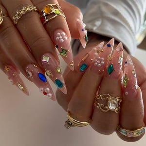 May include: Close-up of long, pointed acrylic nails adorned with colorful gemstones, pearls, and gold accents. Several gold rings with intricate designs and colorful stones are also visible, creating a glamorous aesthetic.