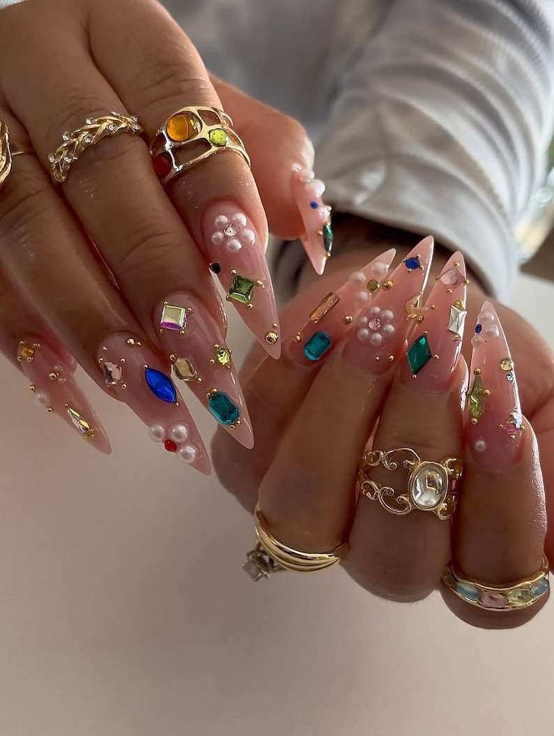 May include: Close-up of long, pointed acrylic nails adorned with colorful gemstones, pearls, and gold accents. Several gold rings with intricate designs and colorful stones are also visible, creating a glamorous aesthetic.