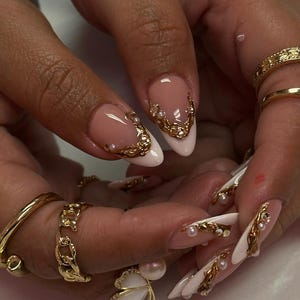 May include: Close-up of manicured nails with a French tip design. The nails are painted white with a pink base and adorned with gold filigree and small pearls. Gold rings are visible on the fingers, including a butterfly ring.