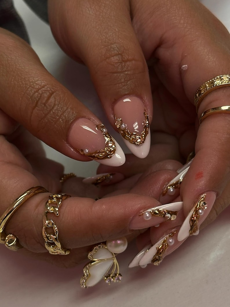 May include: Close-up of manicured nails with a French tip design. The nails are painted white with a pink base and adorned with gold filigree and small pearls. Gold rings are visible on the fingers, including a butterfly ring.