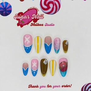 May include: A collection of custom press-on nails with diverse designs. The nails showcase a blend of pink, blue, yellow, and brown, with cartoon artwork and the text "Sugar Nail Nailbox Studio" and "Thank you for your order!"
