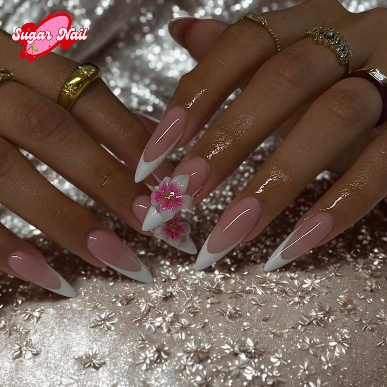 May include: Long, almond-shaped nails with a French manicure design. The nails have a pale pink base with white tips. One nail features a 3D floral embellishment with white petals and pink centers. The image includes the text "Sugar Nail".