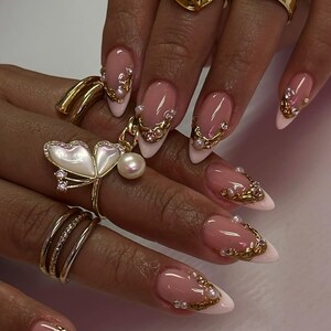 May include: Close-up of a hand with long, almond-shaped nails. The nails feature a pink and white French manicure design, adorned with gold embellishments and small pearls. Several gold rings with intricate designs are also visible.