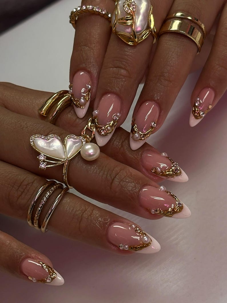 May include: Close-up of a hand with long, almond-shaped nails. The nails feature a pink and white French manicure design, adorned with gold embellishments and small pearls. Several gold rings with intricate designs are also visible.