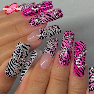 Puede incluir: Uñas largas y cuidadas con un diseño llamativo. Las uñas presentan una combinación de rosa intenso, rayas de cebra negras y estampados de leopardo, acentuadas con brillantes pedrería. El logotipo de Sugar Nail es visible en la esquina superior izquierda.