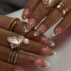 May include: Close-up of a hand with long, almond-shaped nails. The nails feature a pink and white French manicure design, adorned with gold filigree, pearls, and small crystals. Several gold rings, including a butterfly design, are visible.
