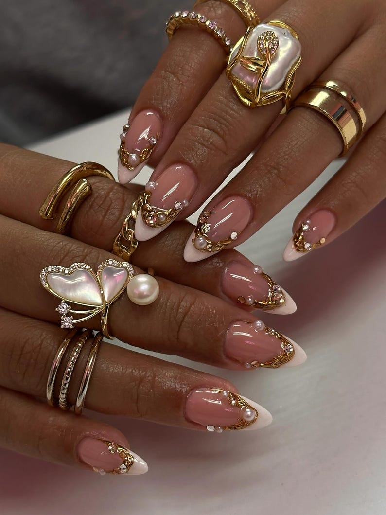 May include: Close-up of a hand with long, almond-shaped nails. The nails feature a pink and white French manicure design, adorned with gold filigree, pearls, and small crystals. Several gold rings, including a butterfly design, are visible.