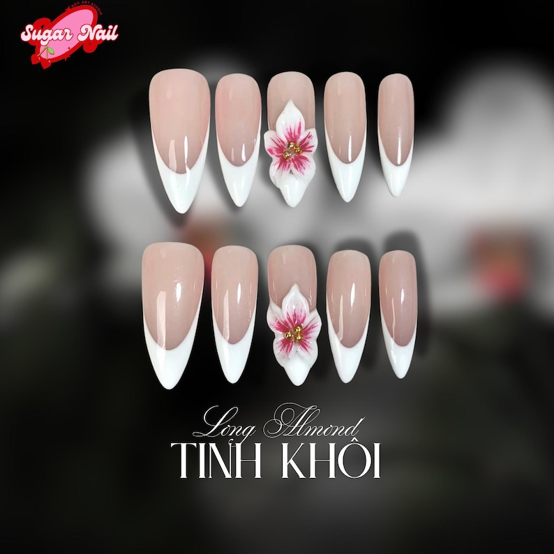 May include: A set of almond-shaped artificial nails with a nude base and white tips. Two nails feature a 3D white flower with pink and gold accents. The text "Long Almond" and "TINH KHOI" is at the bottom.