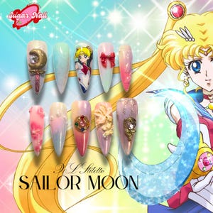 Puede incluir: Un conjunto de diez uñas acrílicas largas en forma de almendra con varios diseños. Las uñas presentan un tema de Sailor Moon, con arte de personajes, lazos, gemas y purpurina. Los logotipos de Sugar Nail y Sailor Moon son visibles.