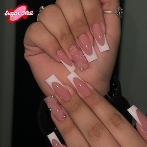 Op de afbeelding: Een set lange, vierkante kunstnagels met een French manicure design. De nagels hebben een lichtroze basis met witte punten. Sommige nagels zijn versierd met 3D bloemversieringen en kleine strass steentjes. Het Sugar Nail logo is zichtbaar.