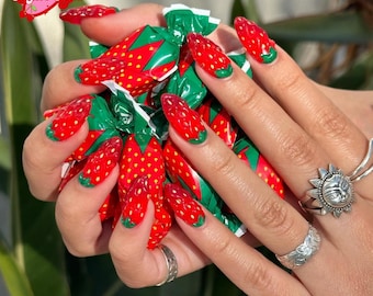 May include: Almond-shaped nails painted with a red strawberry design, featuring white dots and green tips. The image includes wrapped sweets and silver rings. The text "Sugar Nail" is visible in the upper left corner.