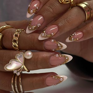 May include: Close-up of a hand with long, almond-shaped nails featuring a French manicure with a white tip and a pink base. The nails are adorned with gold filigree, pearls, and rhinestones. Several gold rings, including a butterfly design, are also visible.