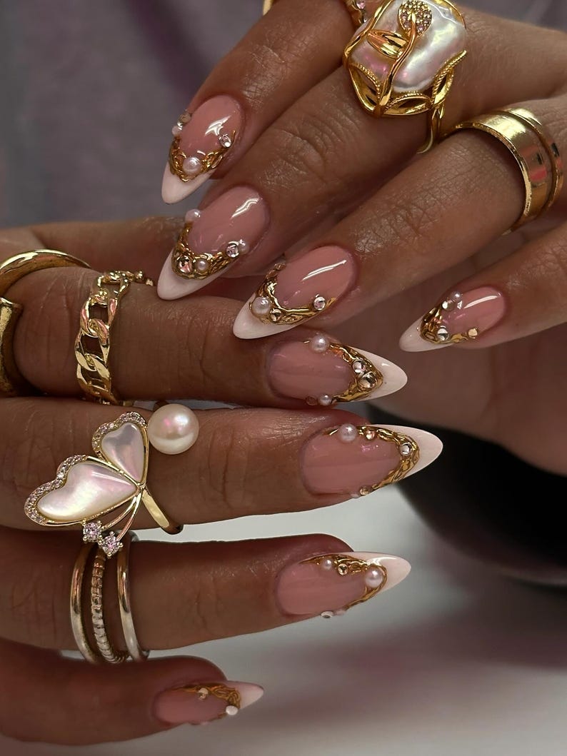 May include: Close-up of a hand with long, almond-shaped nails featuring a French manicure with a white tip and a pink base. The nails are adorned with gold filigree, pearls, and rhinestones. Several gold rings, including a butterfly design, are also visible.