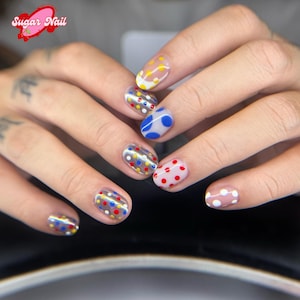 K&ouml;nnte beinhalten: Nahaufnahme einer Hand mit verschiedenen bunten Polka-Dot-Nagelkunst-Designs. Die N&auml;gel zeigen eine Mischung aus Farben, darunter gelbe, rote, blaue und wei&szlig;e Punkte auf einem transparenten oder neutralen Untergrund. Das Sugar Nail-Logo ist oben links sichtbar.
