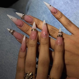 Puede incluir: Uñas largas en forma de estilete con un degradado de rosa a blanco. Cada uña está decorada con pequeños diamantes de imitación transparentes y anillos de metal dorado colocados alrededor de la sección media de la uña, creando un aspecto detallado.