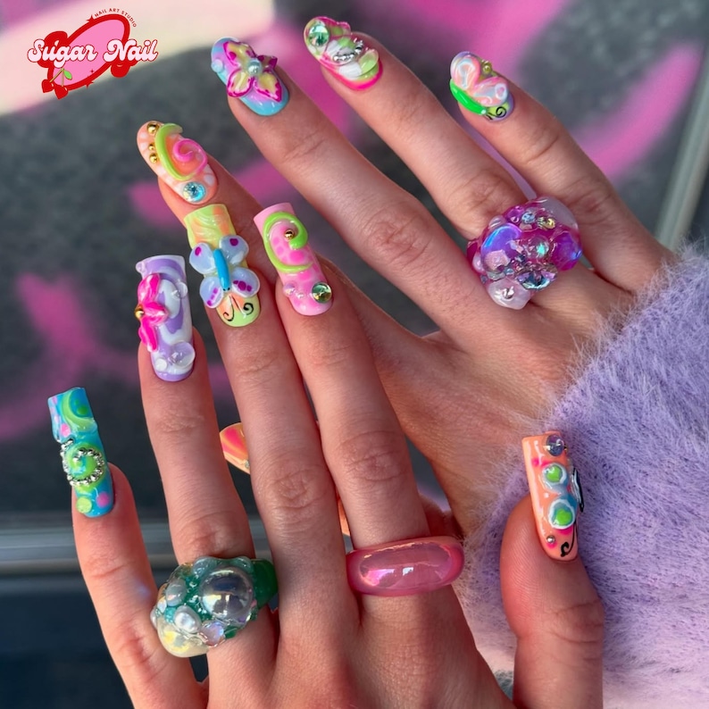 May include: A close-up of a hand with long, colorful, sculpted acrylic nails. The nails feature various designs, including swirls, butterflies, and embellishments. The image also shows several rings with large, decorative stones and a logo that says "Sugar Nail".
