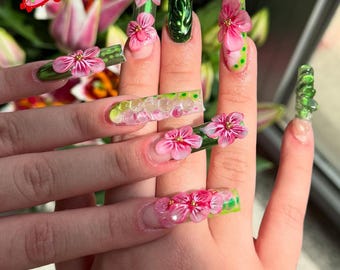 May include: A close-up of a hand with long, decorated fingernails. The nails feature a variety of designs, including 3D pink flowers, green and yellow accents, and a clear, gel-like finish. The logo "Sugar Nail" is visible in the upper left corner.