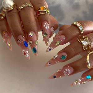 May include: Close-up of long, pointed fingernails decorated with gemstones, pearls, and gold rings. The nails are painted in a nude colour and embellished with colourful gems and small pearls. Gold rings of various designs are also visible.