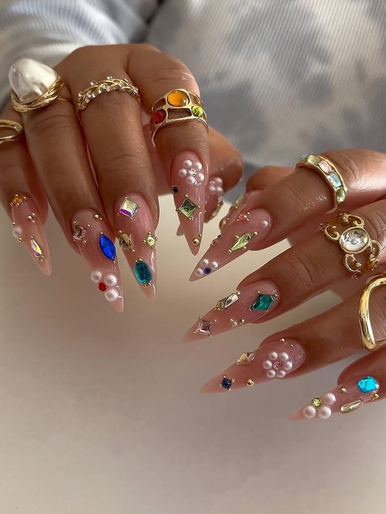 May include: Close-up of long, pointed fingernails adorned with various gemstones, pearls, and gold rings. The nails are painted in a nude color and decorated with an array of colorful gems and small pearls. Gold rings of different designs are also visible.