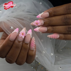 Può includere: Unghie lunghe e appuntite con un design a french manicure rosa e bianco. Ogni unghia presenta un fiore rosa 3D con un centro dorato. Le unghie sono di colore rosa chiaro.