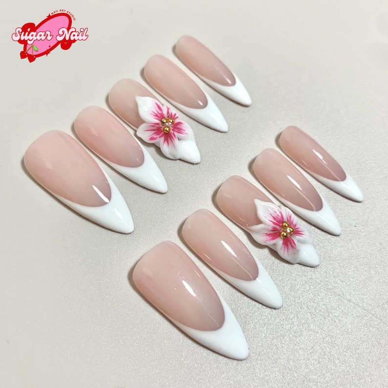 May include: A set of almond-shaped artificial nails with a French manicure design. The nails feature a pale pink base with white tips. Two nails are adorned with 3D white flowers with pink and gold accents. The logo "Sugar Nail" is in the top left corner.