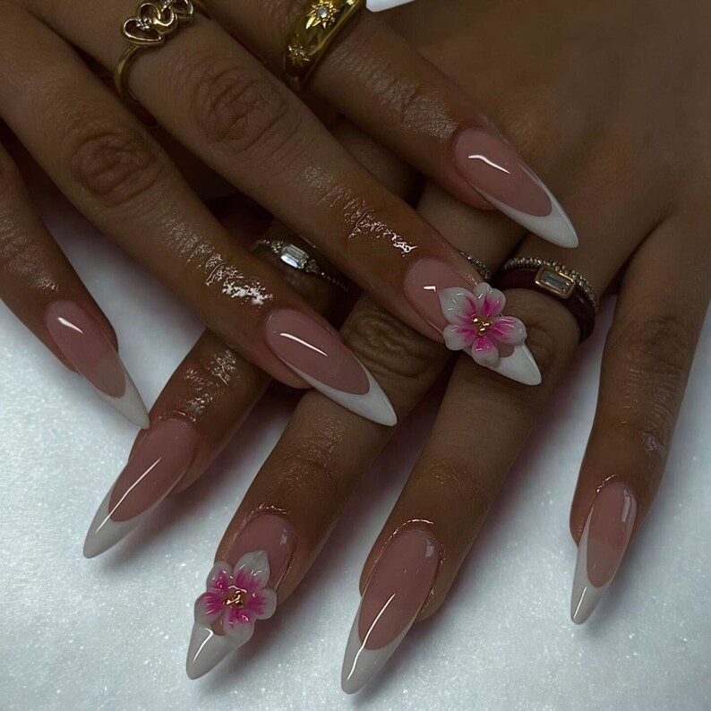May include: Long, almond-shaped nails with a French manicure design. The nails have a nude base with white tips and are adorned with 3D pink and white flower accents. Several gold rings are visible on the fingers.