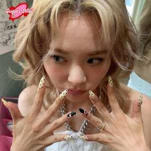 Pode incluir: Close-up das mãos de uma pessoa com unhas longas em forma de amêndoa. As unhas apresentam uma variedade de designs, incluindo bolinhas pretas e brancas, pontas pretas com detalhes dourados e enfeites dourados. A imagem inclui o texto "Sugar Nail".