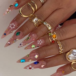 May include: Close-up of a hand with long, pointed, embellished nails. The nails are adorned with colorful gems, pearls, and gold accents. Several gold rings of various designs, including a square diamond ring, are worn on the fingers.