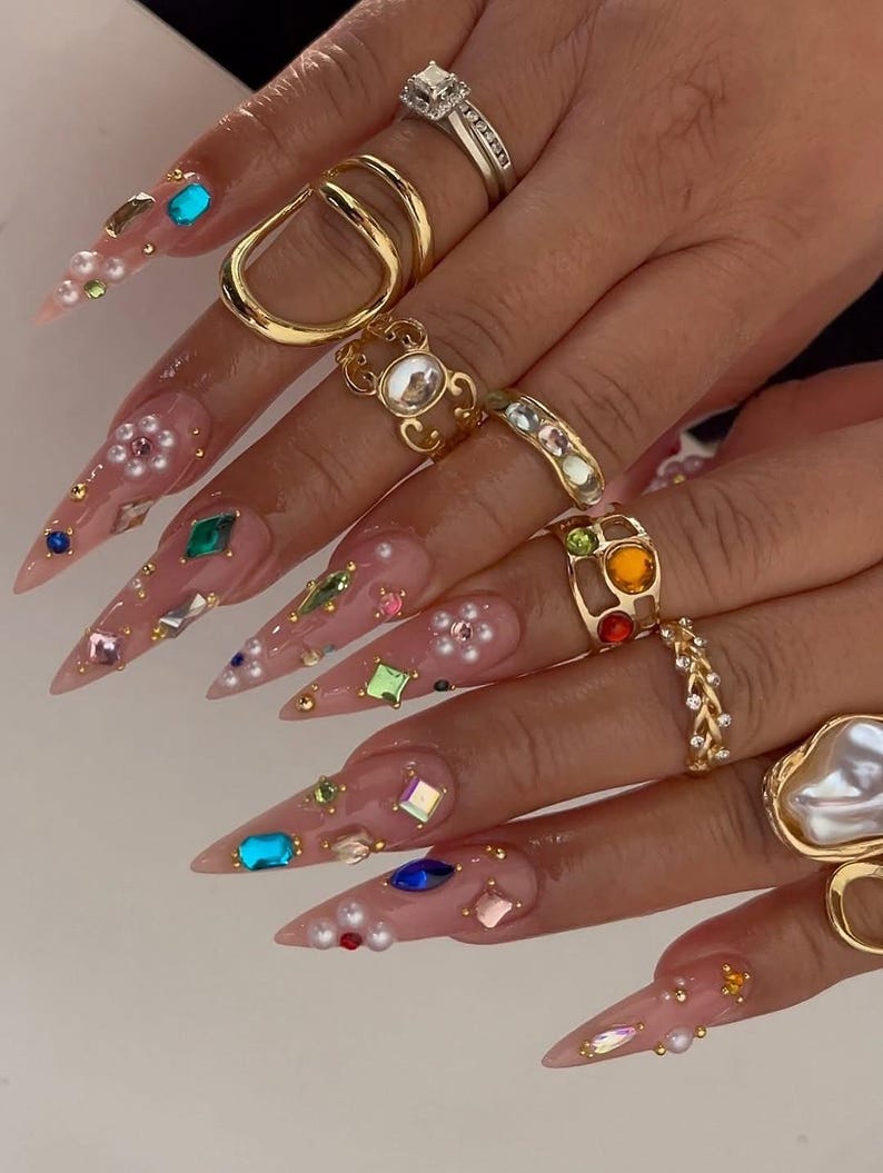 May include: Close-up of a hand with long, pointed, embellished nails. The nails are adorned with colorful gems, pearls, and gold accents. Several gold rings of various designs, including a square diamond ring, are worn on the fingers.
