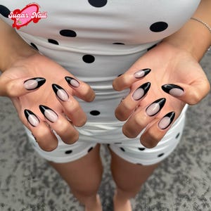 May include: Close-up of hands with almond-shaped nails. The nails feature a black French tip design with a nude base. The person is wearing a white polka dot outfit. The Sugar Nail logo is visible in the upper left corner.