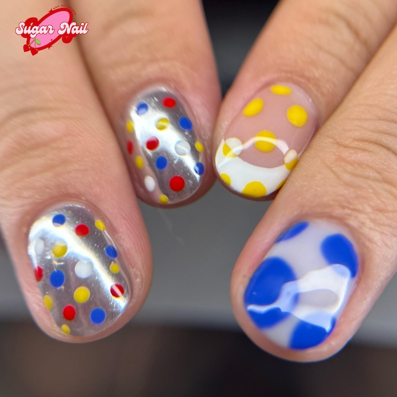 K&ouml;nnte beinhalten: Nahaufnahme von manik&uuml;rten N&auml;geln mit bunten Polka-Dot-Designs. Zwei N&auml;gel haben ein silbernes Chrom-Finish mit roten, gelben, blauen und wei&szlig;en Punkten. Andere N&auml;gel zeigen gelbe und blaue Polka-Dots auf klarer Basis.