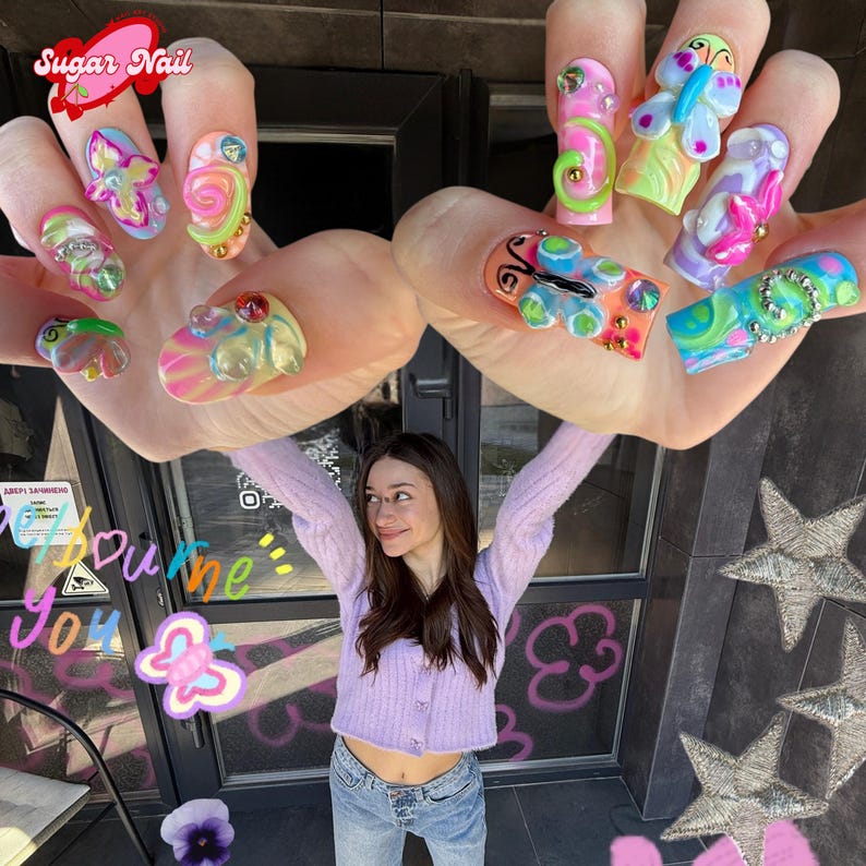 May include: A person holding up hands with colorful, artistic nail designs. The nails feature 3D butterfly, swirl, and gem embellishments in shades of pink, green, blue, and yellow. The text "Sugar Nail" is visible in the upper left corner.