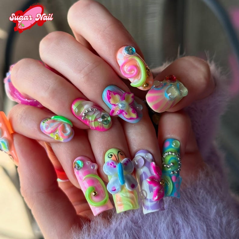 May include: A hand displaying long, colorful, sculpted acrylic nails. Each nail features a unique design with vibrant colors, including pink, green, yellow, and blue, adorned with 3D embellishments, gems, and swirls. The text "Sugar Nail" is visible.