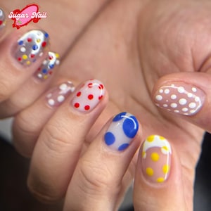 K&ouml;nnte beinhalten: Eine Hand mit einer Manik&uuml;re mit verschiedenen Polka-Dot-Designs. Die N&auml;gel sind mit klarem, rosa, blauem und gelbem Nagellack bemalt, jeder mit verschiedenfarbigen Polka-Dots verziert. Das Sugar Nail-Logo ist oben links sichtbar.