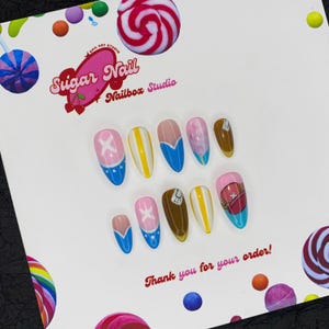 May include: A collection of ten false nails with diverse designs in pink, blue, yellow, and brown. The nails display patterns such as an X, stripes, and a cartoon character. The text "Sugar Nail Nailbox Studio" and "Thank you for your order!" are also visible.