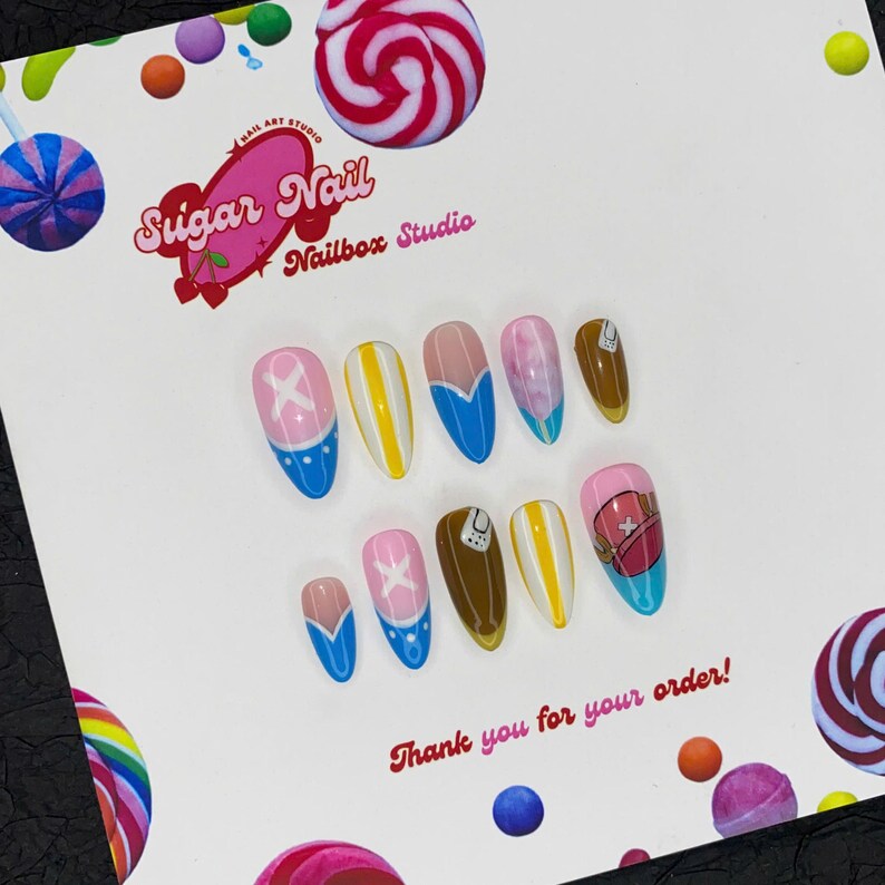 May include: A collection of ten false nails with diverse designs in pink, blue, yellow, and brown. The nails display patterns such as an X, stripes, and a cartoon character. The text "Sugar Nail Nailbox Studio" and "Thank you for your order!" are also visible.