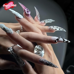 Puede incluir: Una mano que muestra uñas acrílicas largas y puntiagudas con varios diseños. Algunas uñas presentan diseños florales en 3D, mientras que otras muestran patrones como lunares, espirales y detalles con tachuelas. Las uñas son en tonos rosa, blanco, negro y transparente.